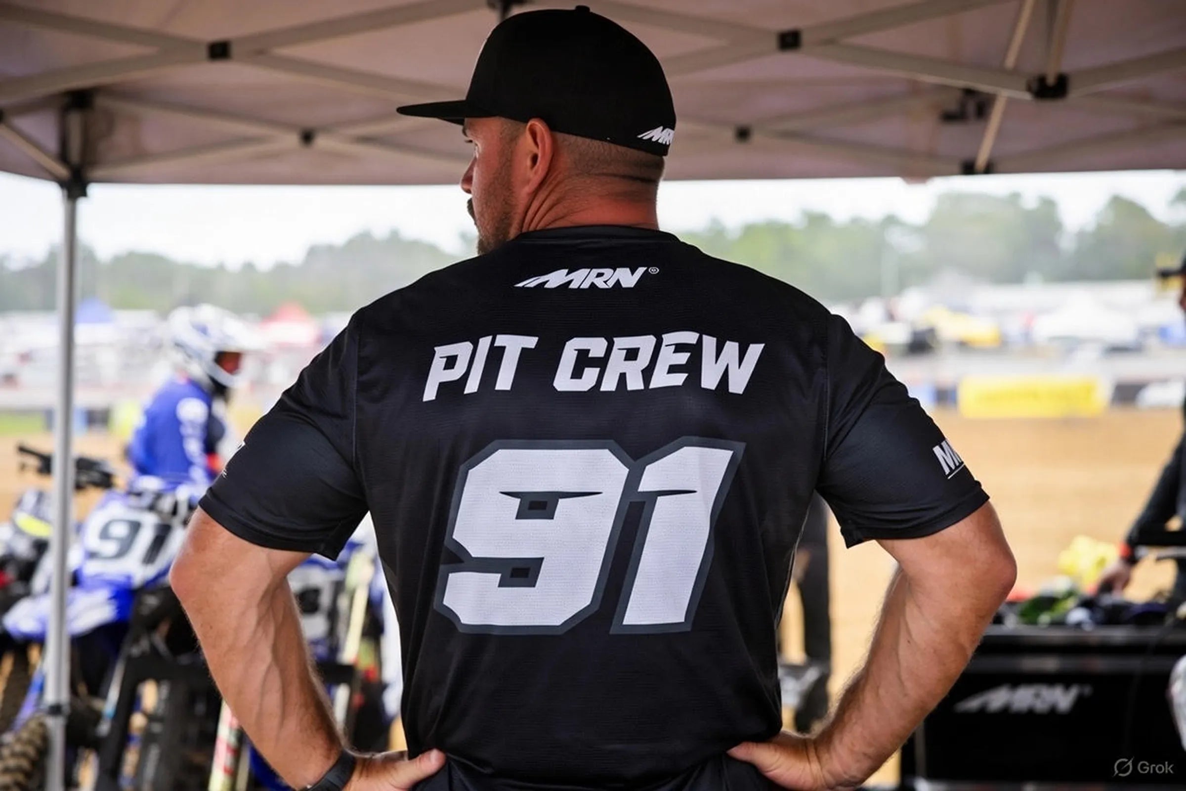 Custom motocross pit crew shirt with race number on the back, worn by a crew member standing under a canopy at the track
