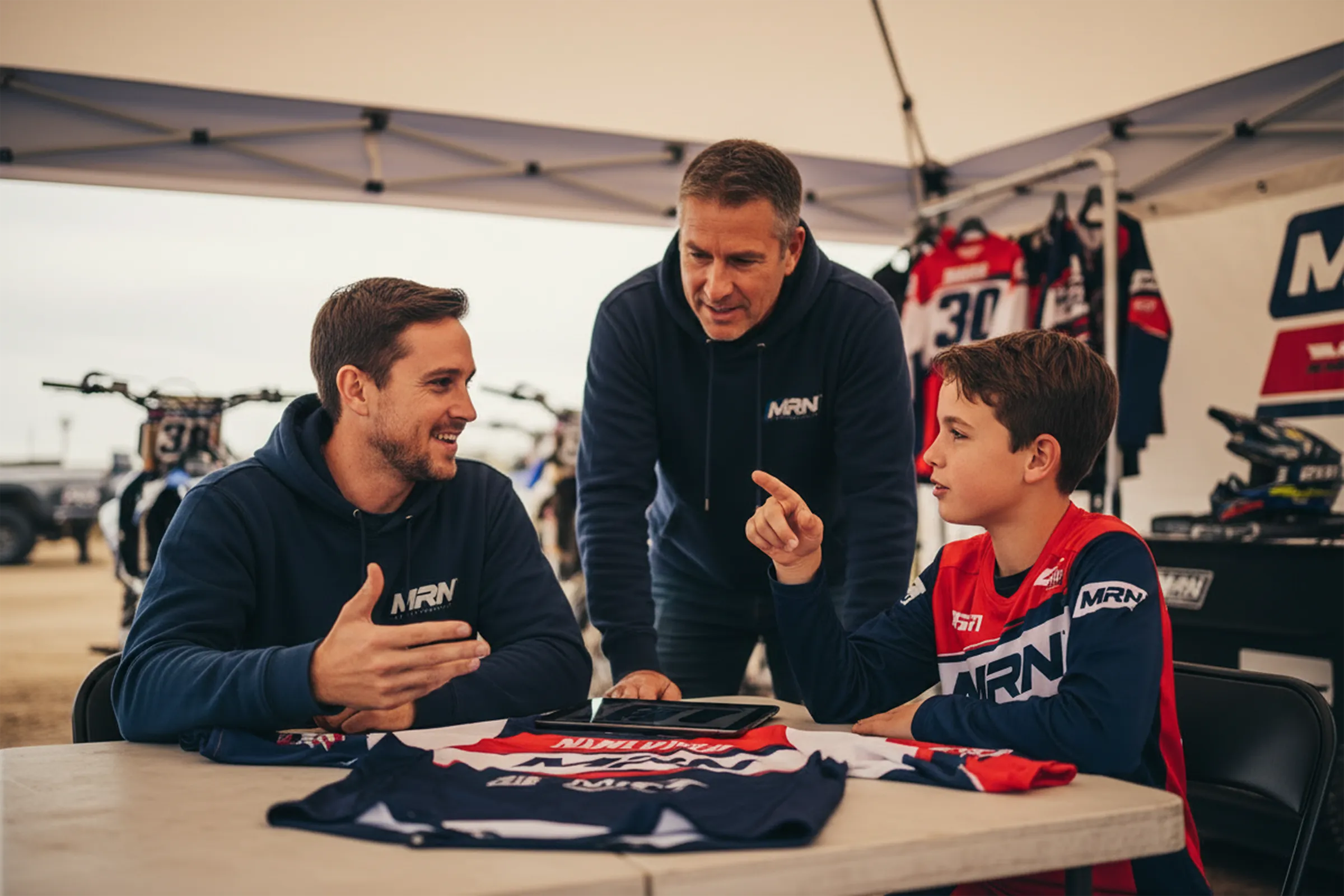 MRN graphic designer collaborating with a motocross rider and parent to design a custom motocross jersey using a tablet at the track